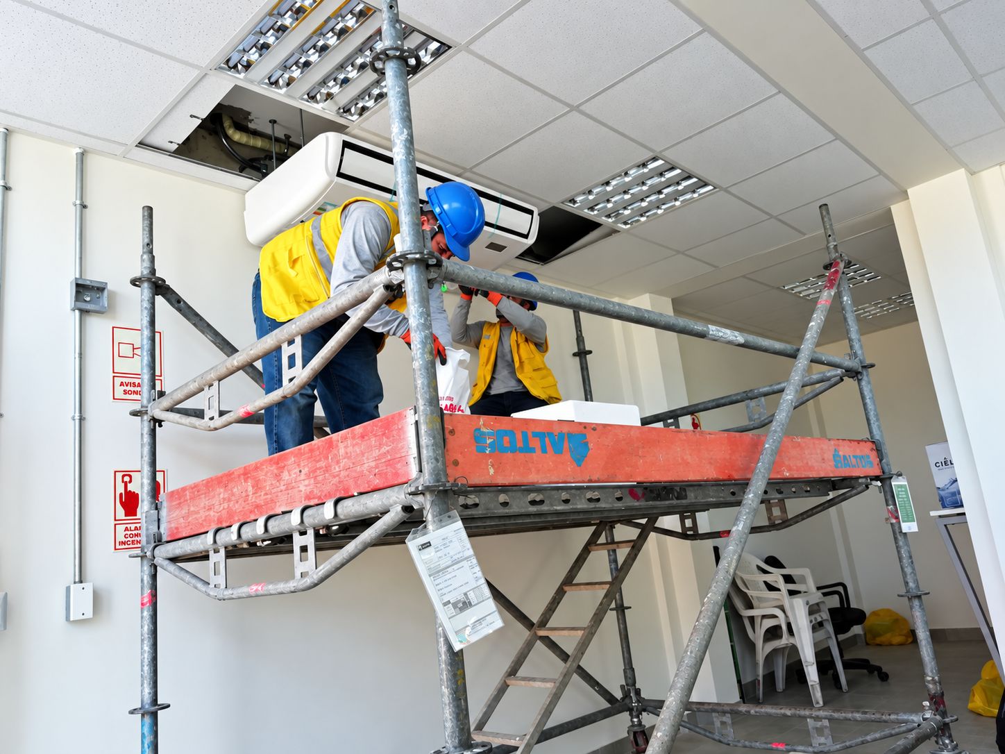 Dos técnicos con casco y chaleco sobre un andamio instalando un equipo de aire acondicionado en el techo de una oficina.